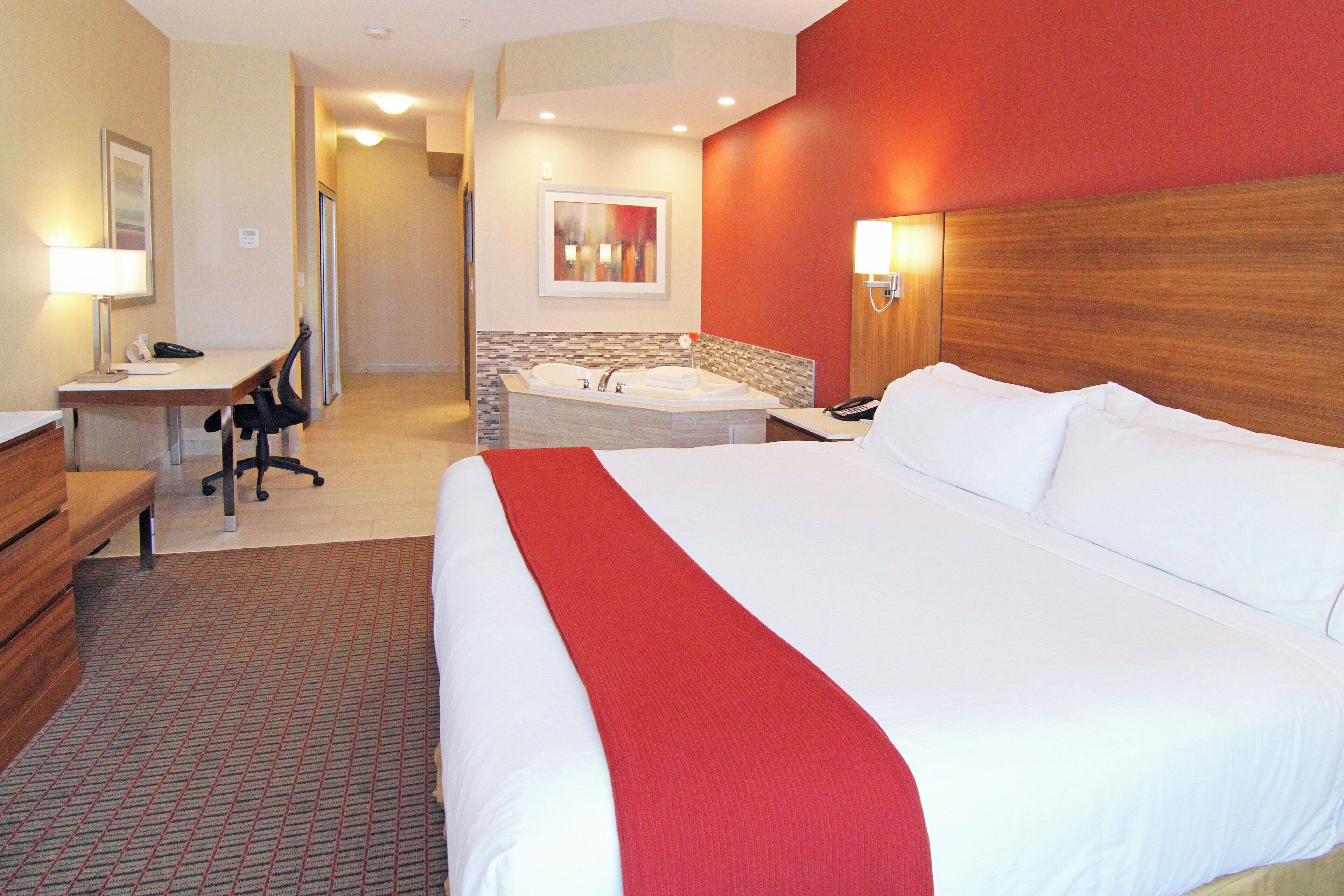 A hotel room with a large bed, a desk with a chair, a small sitting area, a bathroom with a bathtub, and a red accent wall.