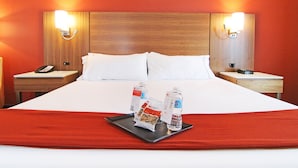 A hotel room with a large bed, two bedside tables, a nightstand with a phone, and a tray with bottled water and snacks.