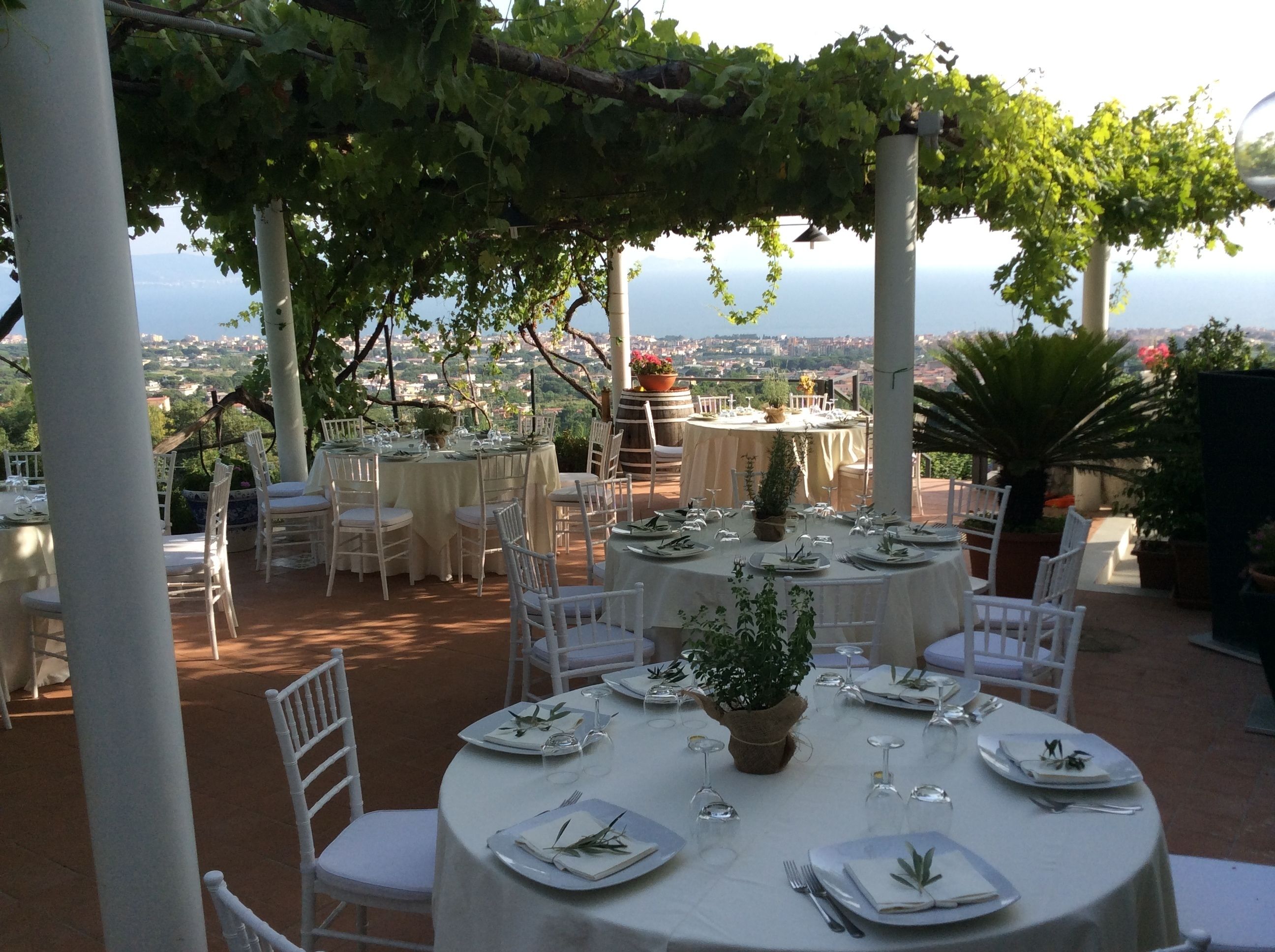 Un área de comedor al aire libre con mesas redondas dispuestas para una comida, rodeada de sillas blancas y con vistas panorámicas a la ciudad y al mar.