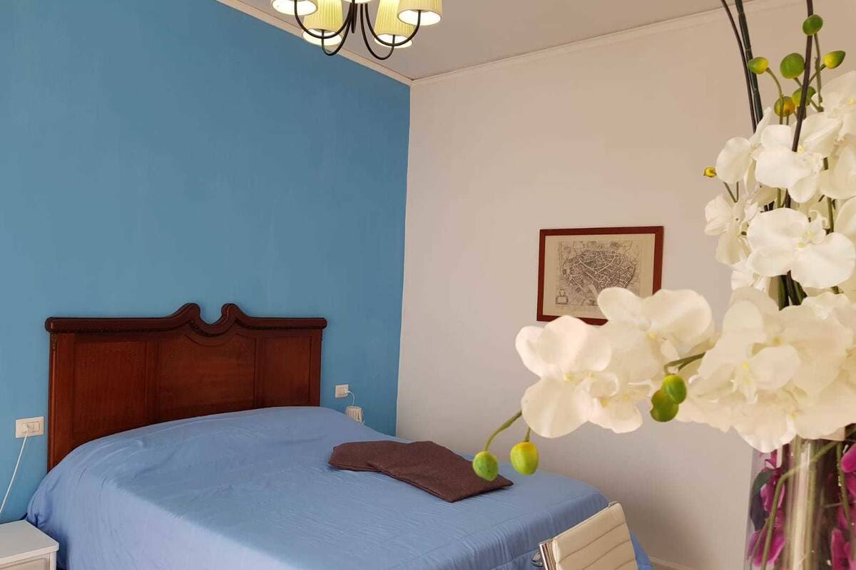 A bedroom with a blue wall, a wooden bed frame, a blue bedspread, a brown pillow, a white nightstand, a white chair, a vase with white flowers, and a framed picture on the wall.