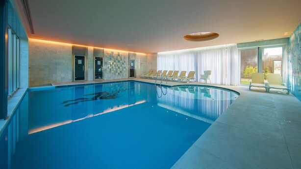 A large indoor swimming pool with clear blue water, surrounded by lounge chairs and a modern ceiling design. - HD Duecitânia Design Hotel (Penela)