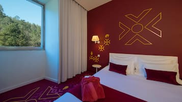 A hotel room with a large bed, a window with curtains, a wall with geometric patterns, and a small table with a lamp.
