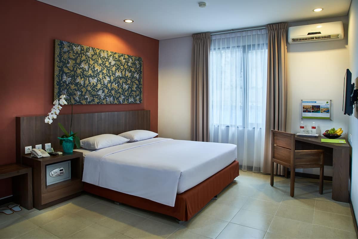 Habitación de hotel con cama, escritorio, silla, teléfono, caja fuerte, planta y un cuadro en la pared.