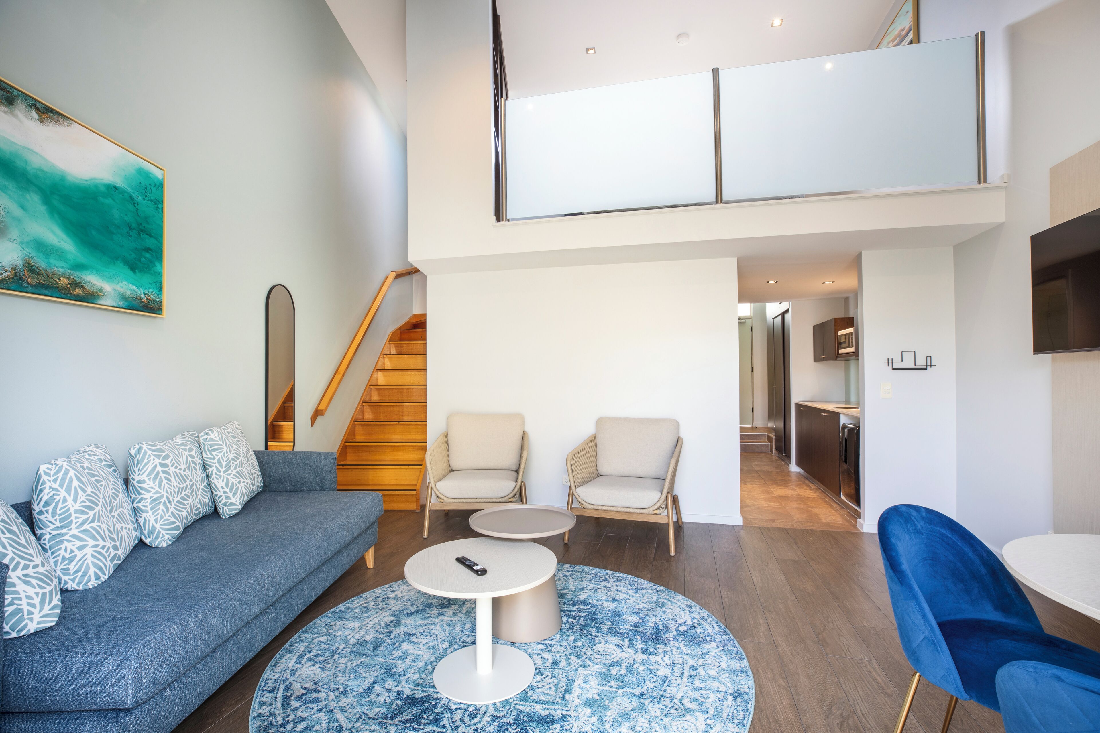 Ein modernes Wohnzimmer mit einem grauen Sofa, zwei Sesseln, einem Couchtisch und einem blauen Teppich. Eine Treppe führt zu einer oberen Ebene.