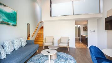 Ein modernes Wohnzimmer mit einem grauen Sofa, zwei Sesseln, einem Couchtisch und einem blauen Teppich. Eine Treppe führt zu einer oberen Ebene.