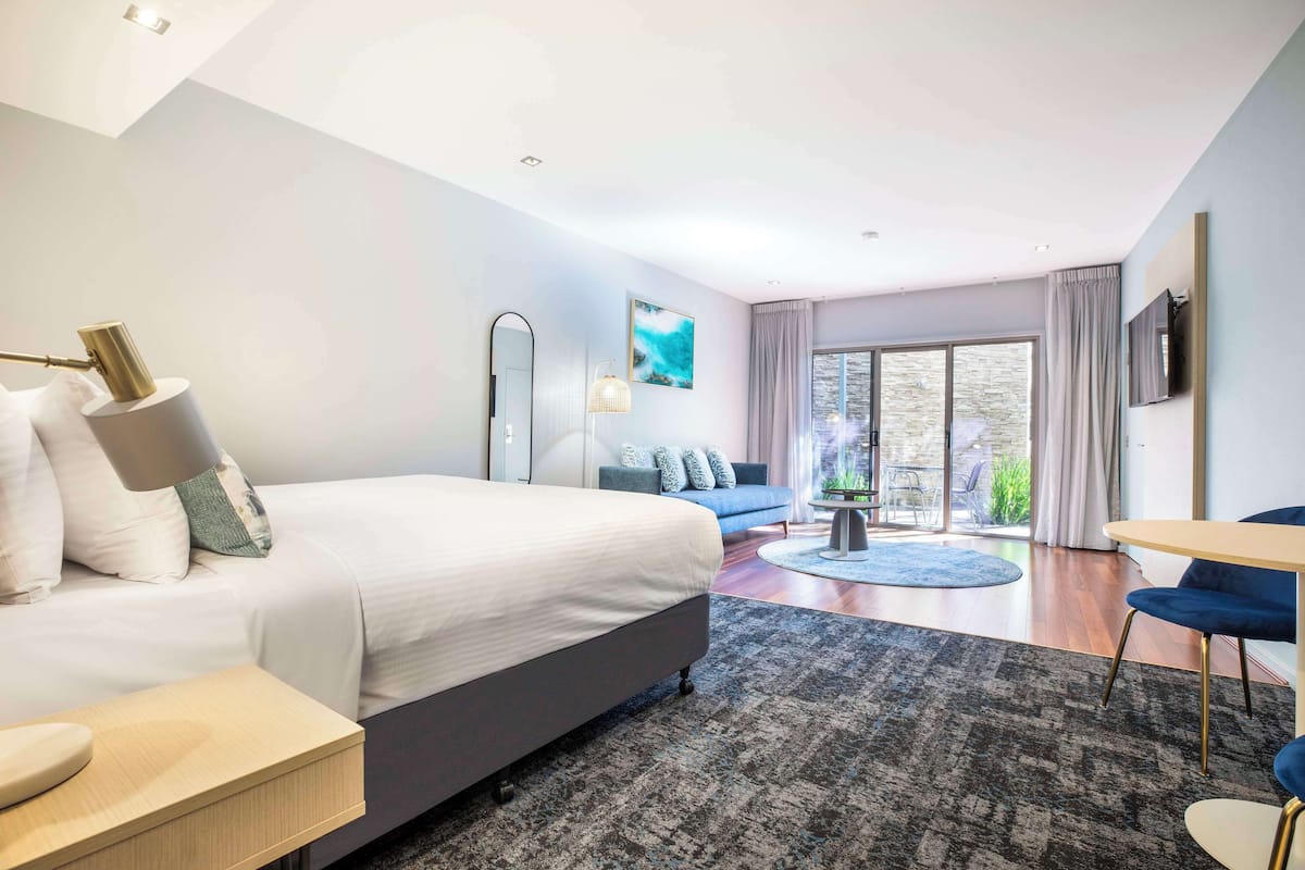 A modern hotel room with a large bed, a seating area with a blue sofa, a small round table, and a large window offering a view of the outdoors.