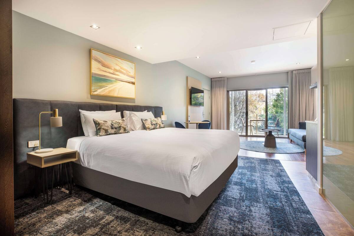 A modern bedroom with a large bed, a bedside table, a lamp, and a view of the outdoors through a glass door.