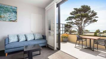 Un balcón con sofá, mesa y sillas, una puerta corredera y un ventanal grande con vista a un árbol.
