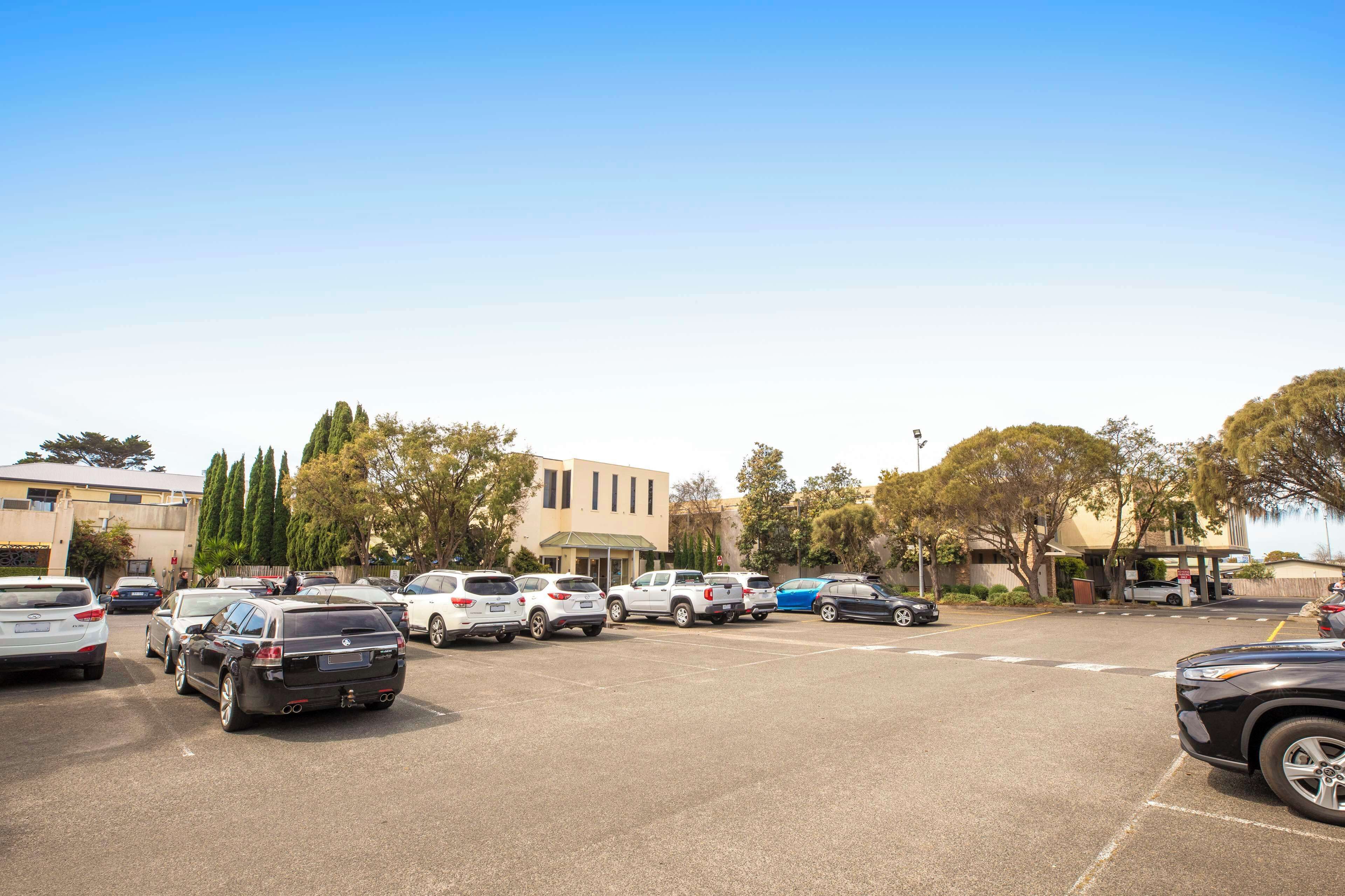 Un estacionamiento con varios automóviles y un edificio de techo plano con múltiples ventanas.