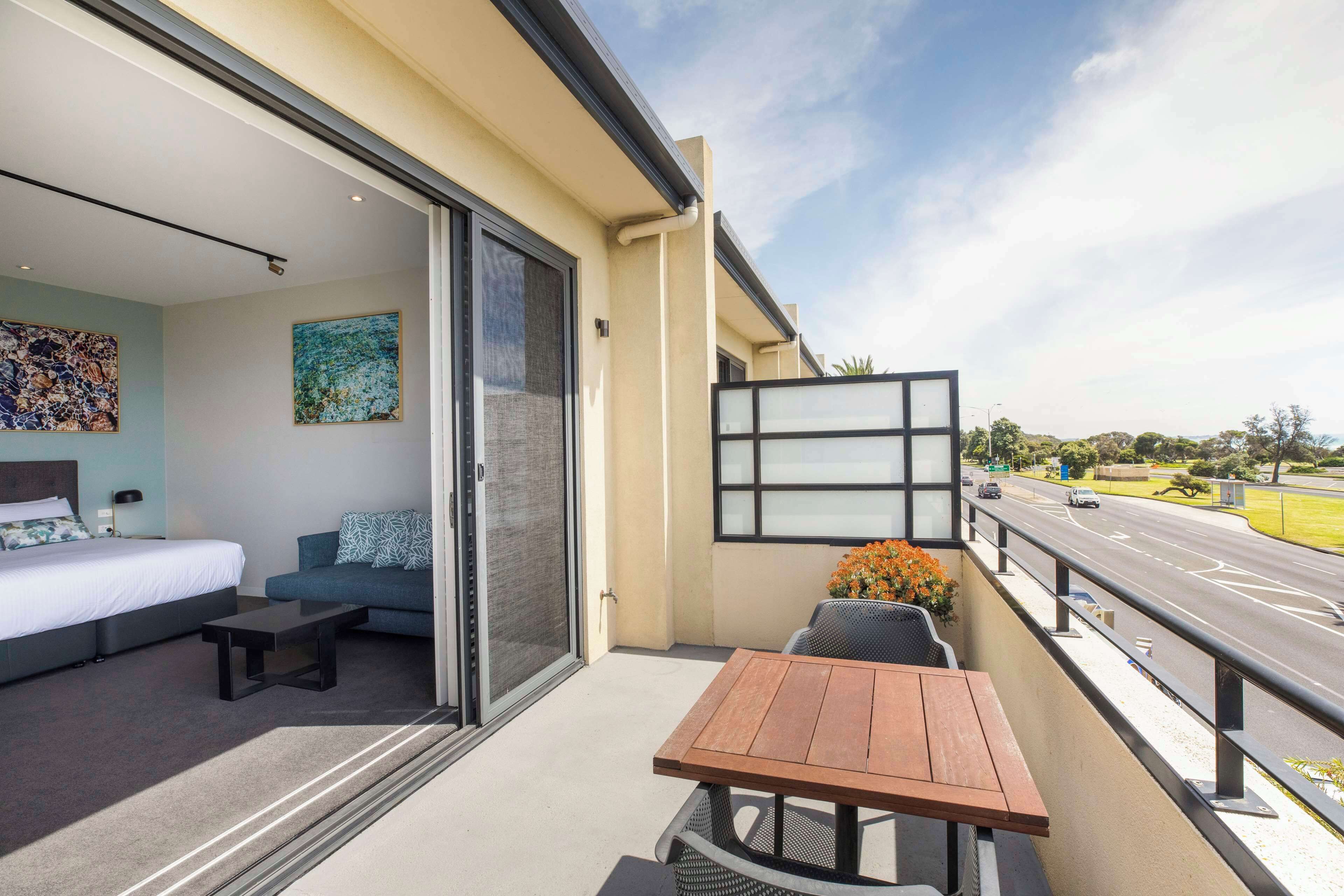 A balcony with a wooden table and chairs, a sofa, a bed, and a large window overlooking a road.