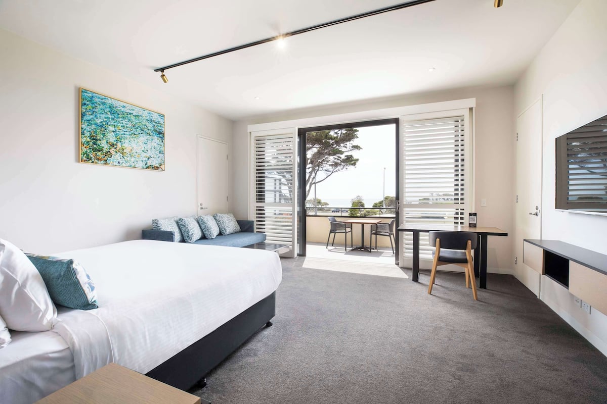 A modern bedroom with a large bed, a desk, and a balcony with a view.