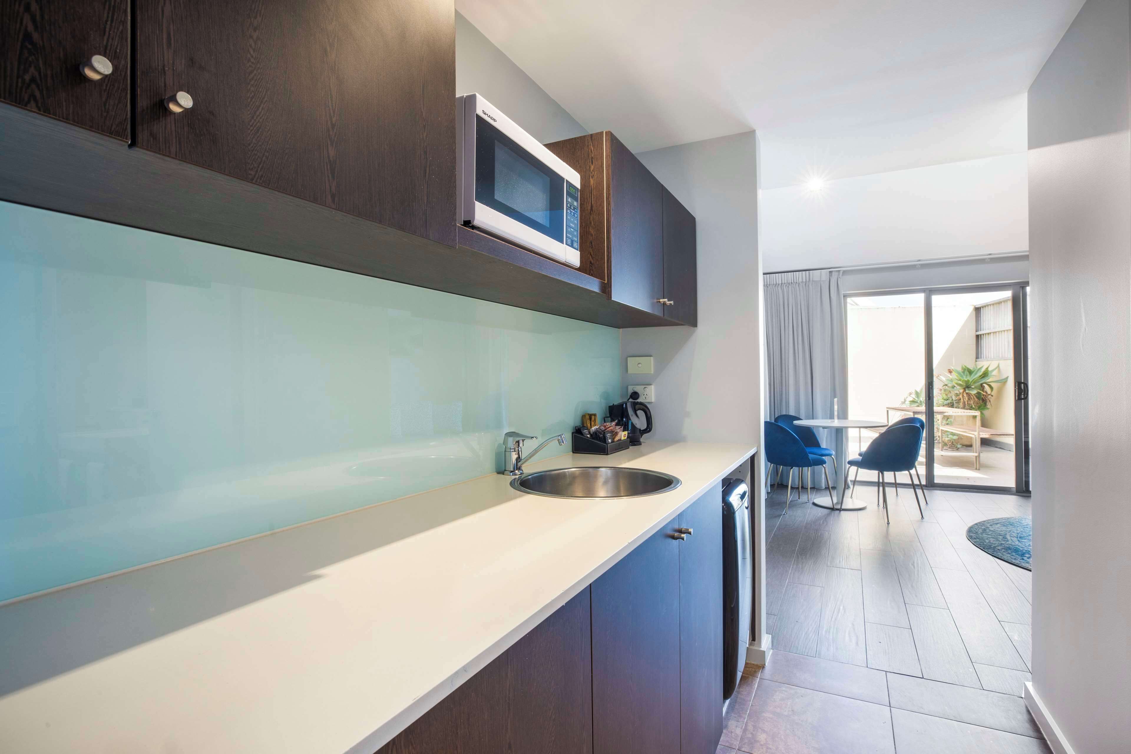 Una cocina moderna con microondas integrado, un lavamanos y armarios. Se observa un comedor con una mesa y sillas a través de una puerta de vidrio.