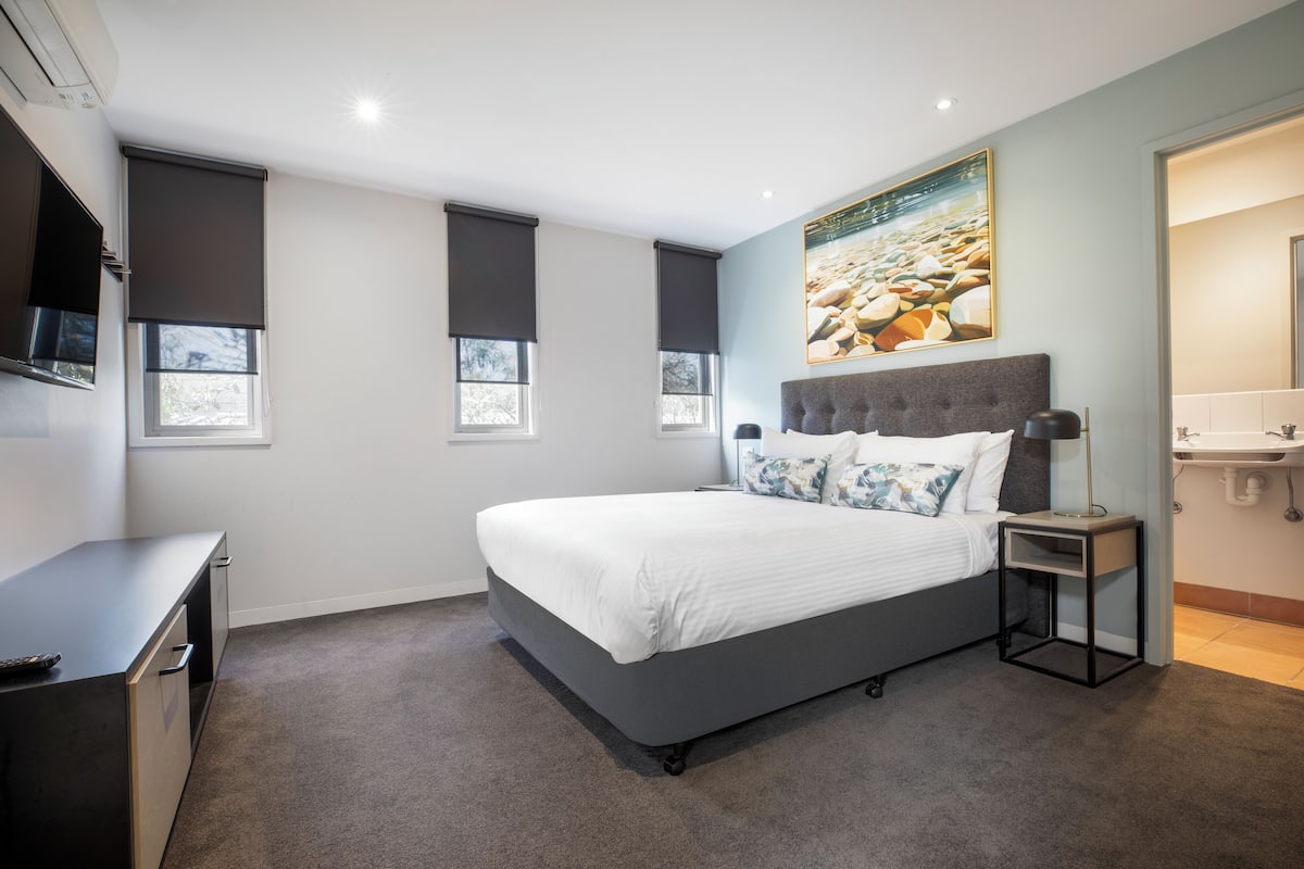 A bedroom with a bed, a bedside table, a TV, and a bathroom with a sink.