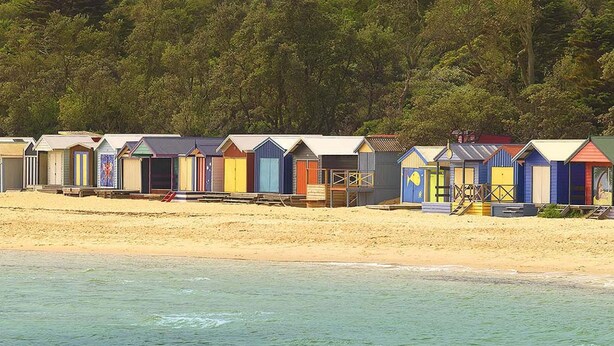 A row of beach huts with a sandy beach and water in the foreground.