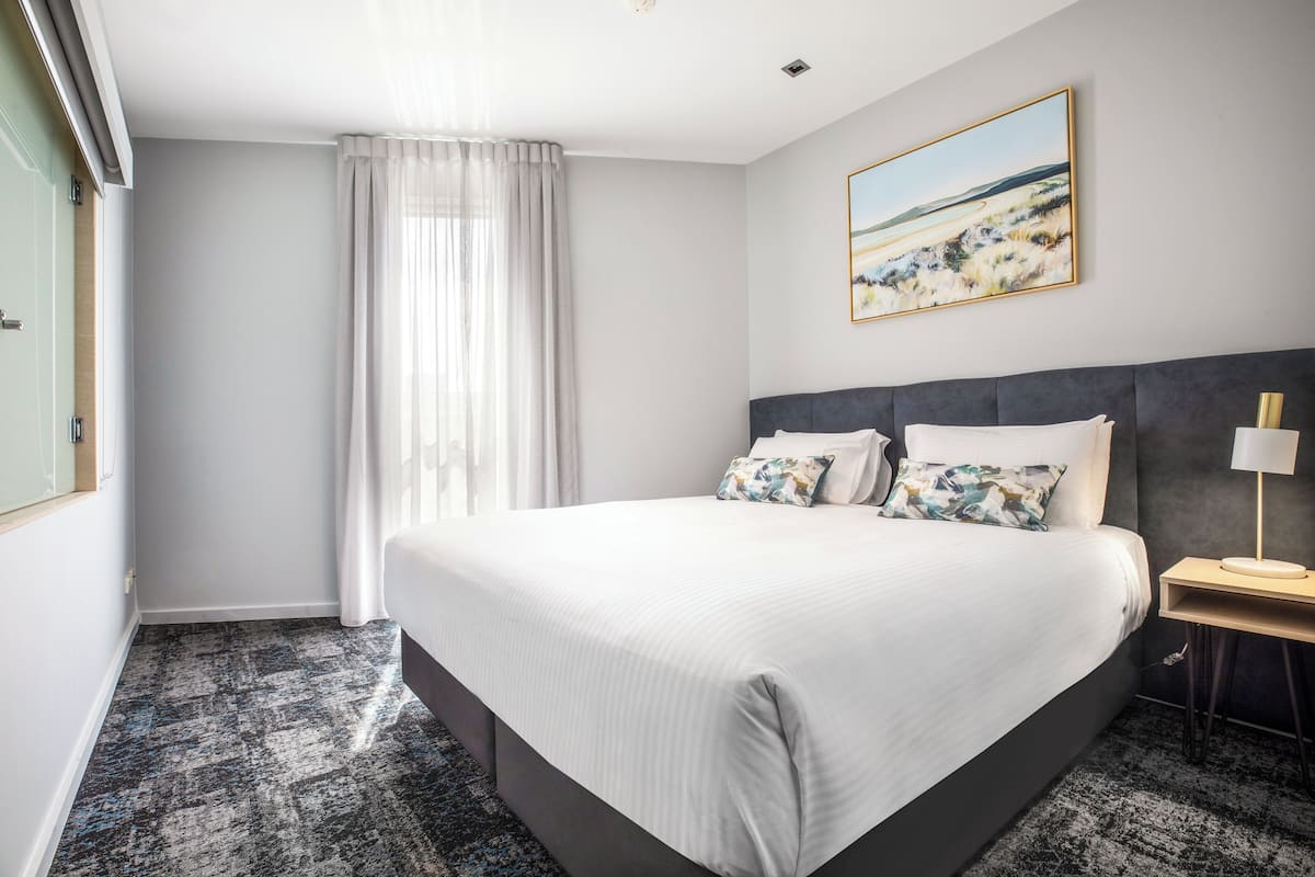 A hotel room with a large bed, a bedside table with a lamp, a painting on the wall, and a window with curtains.