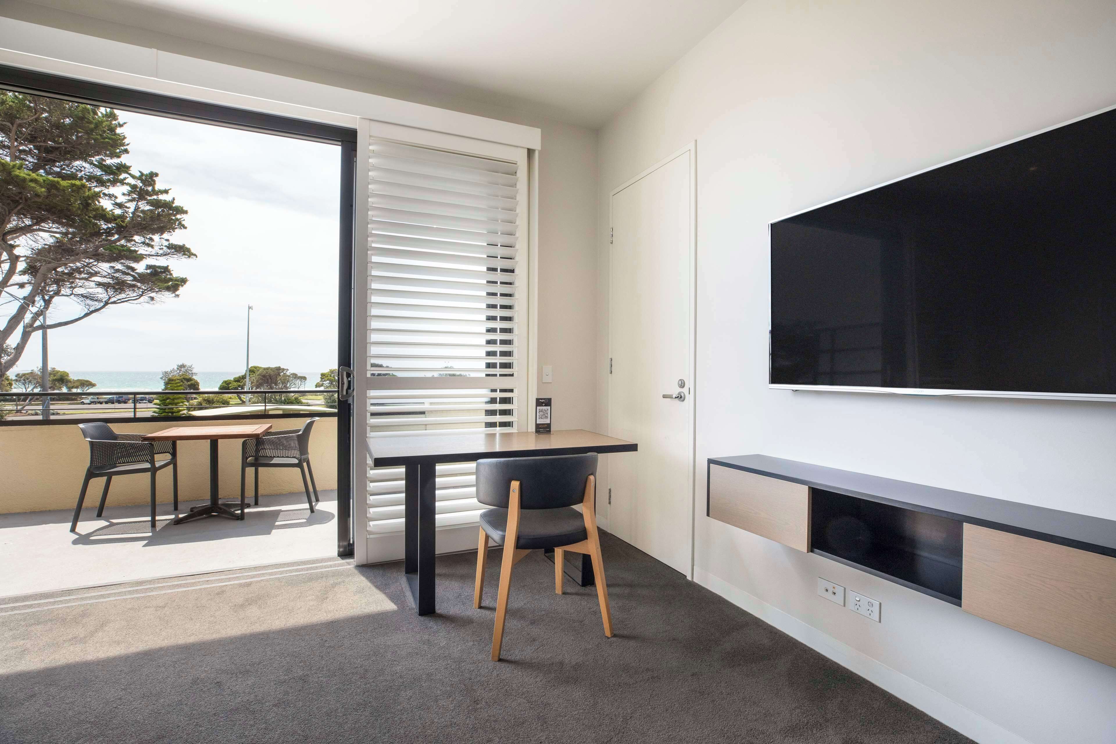 A modern living room with a large TV mounted on the wall, a dining area with a table and chairs, and a view of the outdoors through a sliding door.