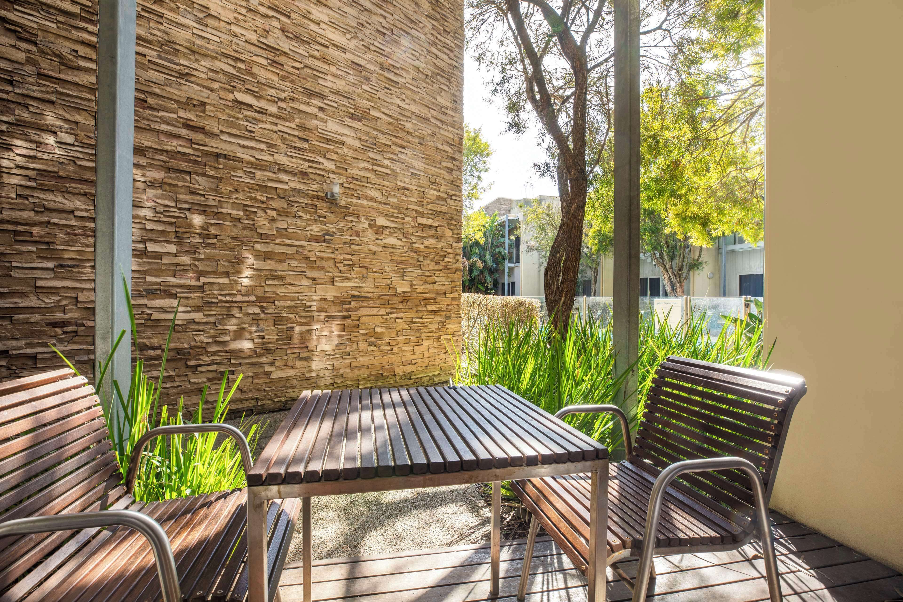 Un patio al aire libre con una mesa de madera y bancos, rodeado por un muro de piedra y follaje.