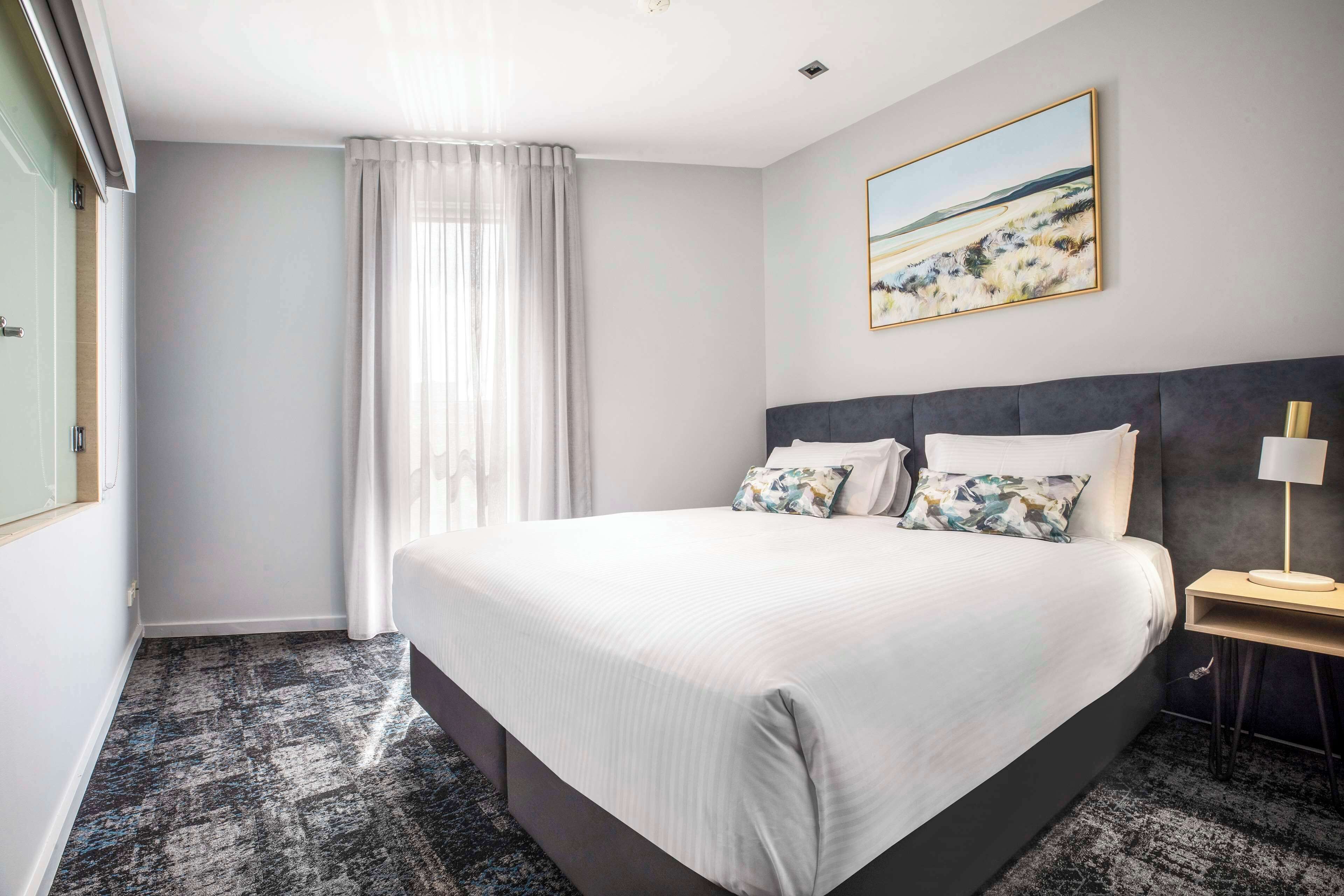 A hotel room with a large bed, a bedside table with a lamp, a painting on the wall, and a window with curtains.