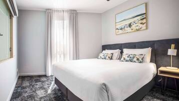Habitación de hotel con una cama grande, una mesita de noche con lámpara, un cuadro en la pared y una ventana con cortinas.