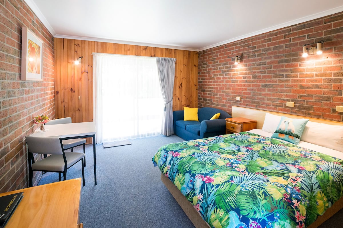 Ein Zimmer mit einem Bett, einem Schreibtisch und einem Stuhl. Es gibt ein Fenster mit Vorhängen und eine Ziegelwand.