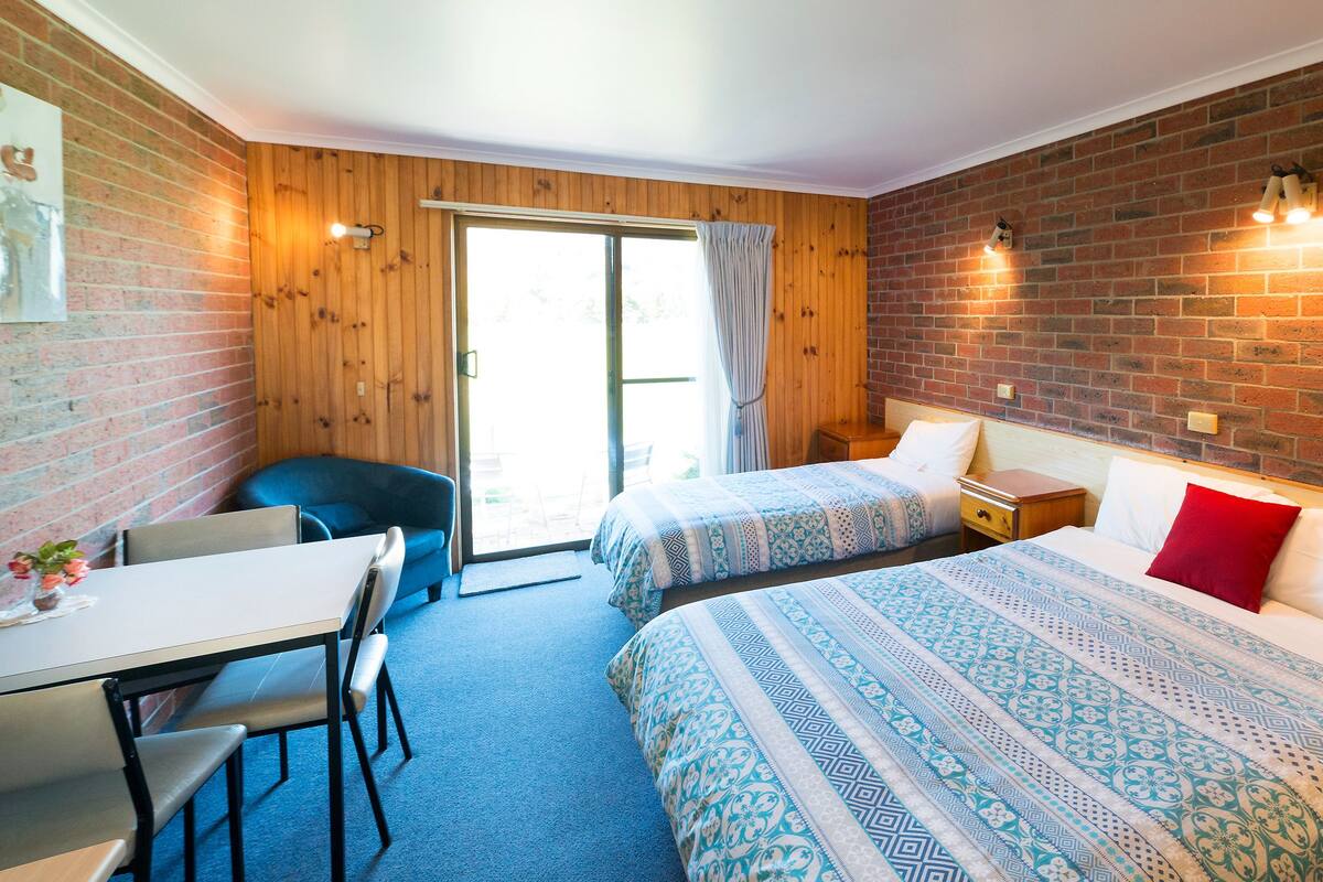 Ein Zimmer mit zwei Betten, einem Schreibtisch und einem Stuhl. Eine Ziegelwand und eine Holztür sind zu sehen.