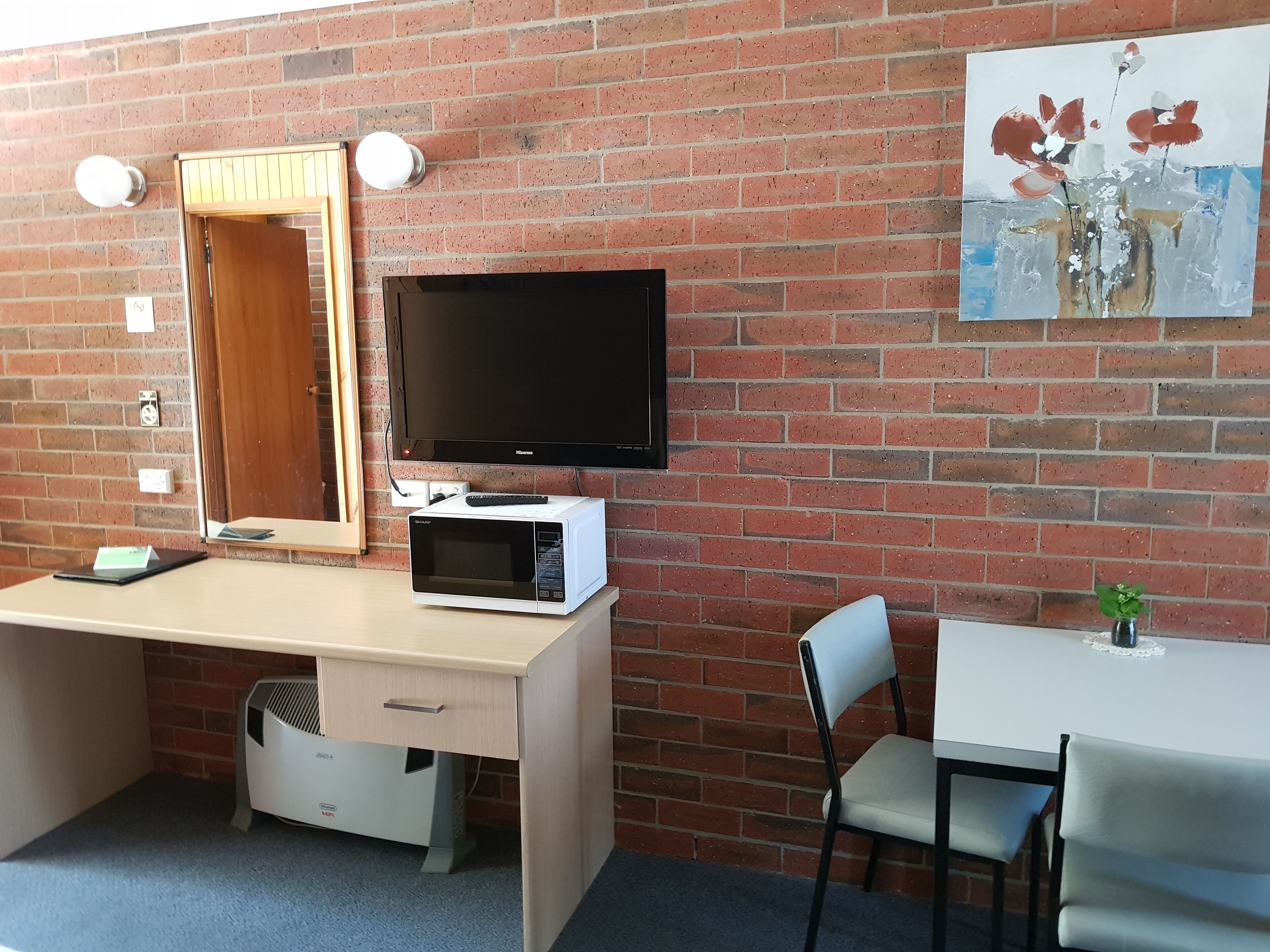 A room with a brick wall, a flat-screen TV, a microwave, a desk, a chair, and a small table with a plant.