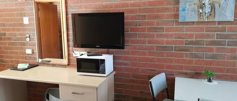 A room with a brick wall, a flat-screen TV, a microwave, a desk, a chair, and a small table with a plant.