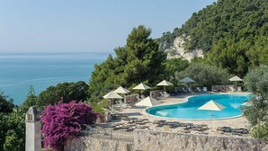 A pool area with lounge chairs and umbrellas, surrounded by greenery and overlooking a body of water. - Baia Delle Zagare - Handwritten Collection (Mattinata)
