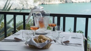 A table set with a pitcher, glasses, and a basket of bread on a balcony with a view of a rocky coastline. - Baia Delle Zagare - Handwritten Collection (Mattinata)