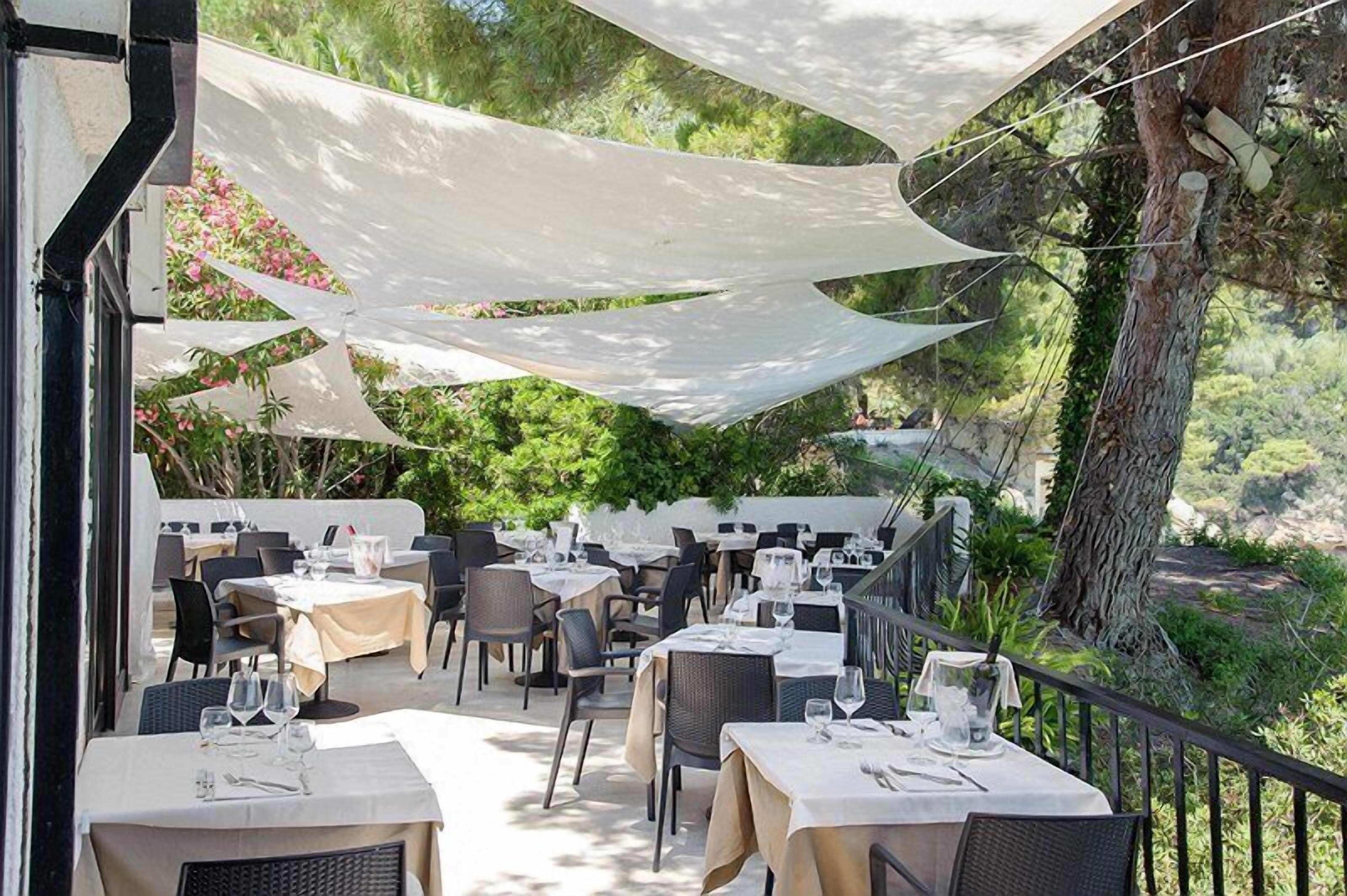 An outdoor dining area with tables and chairs, shaded by white canopies, set against a backdrop of greenery and trees.