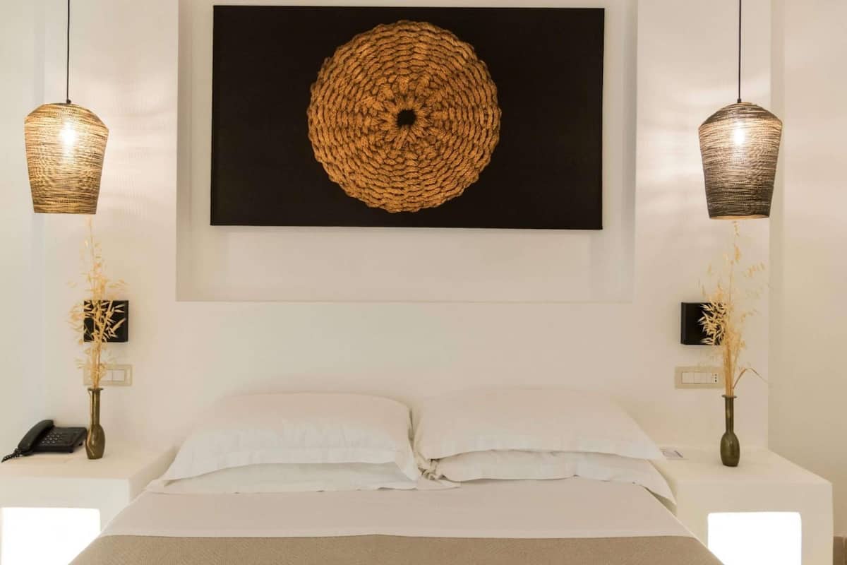 Una cama con sábanas blancas, dos mesitas de noche con lámparas, una obra de arte en la pared y un interruptor.