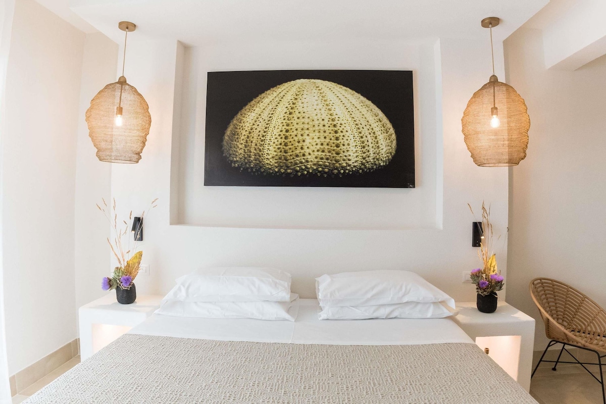 Un dormitorio con una cama, dos mesitas de noche, una silla, una obra de arte en la pared y dos lámparas colgantes.