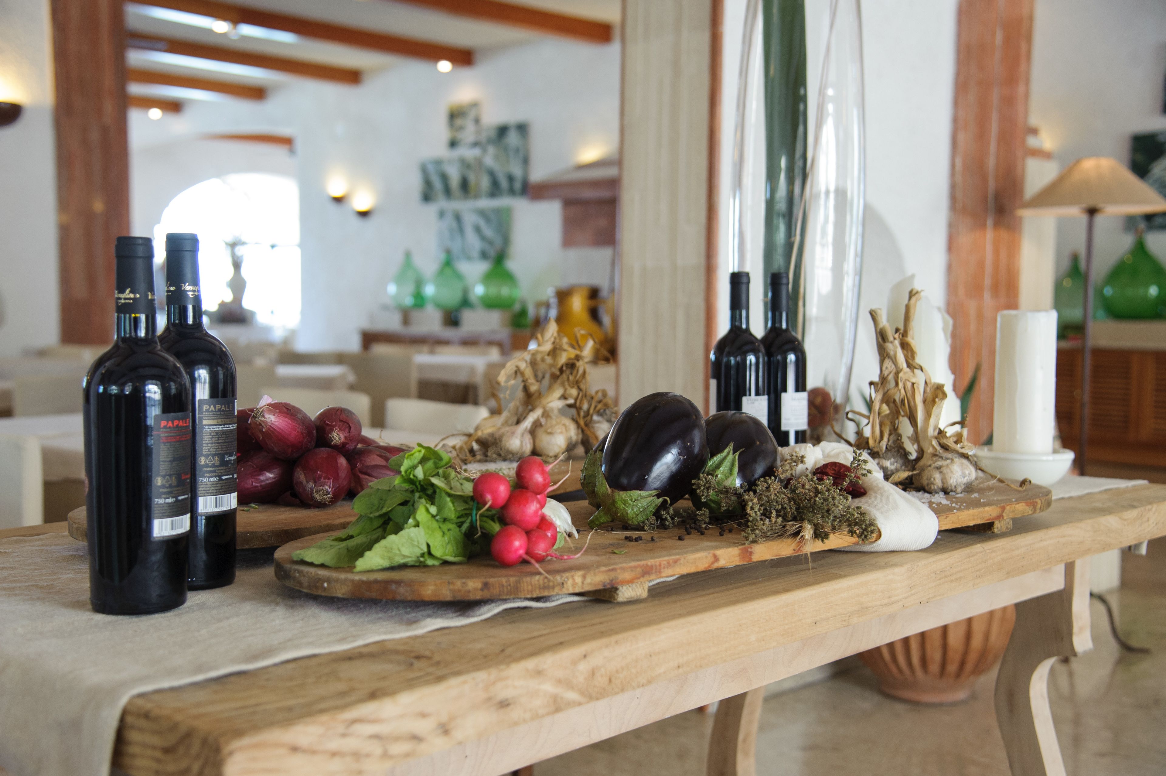 A wooden table with wine bottles, fresh vegetables, and decorative items in a well-lit room with wooden beams and a modern interior.