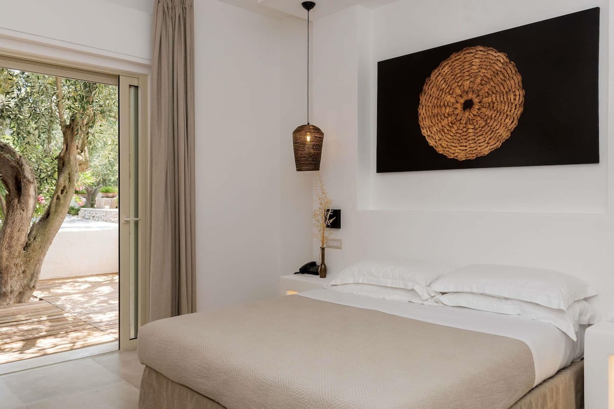 Un dormitorio con una cama grande, una obra de arte en la pared, una mesita de noche con una lámpara y vista a un árbol a través de una puerta corrediza.