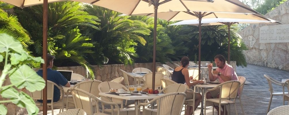 An outdoor dining area with wicker chairs and tables, surrounded by lush greenery and a stone wall.