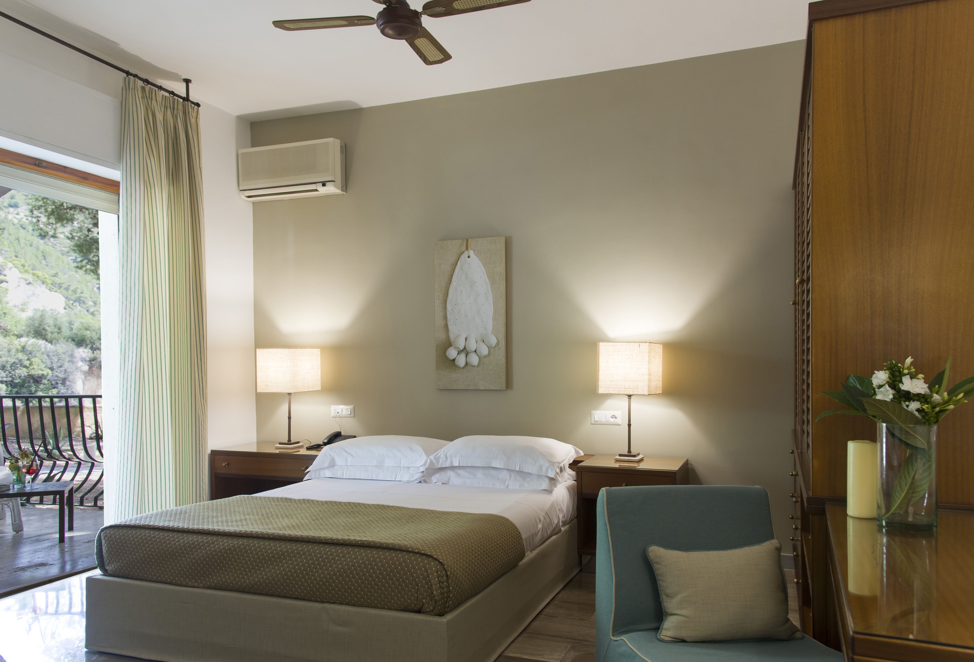 A hotel room with a bed, two bedside tables with lamps, a chair, a desk with a vase, and a ceiling fan.