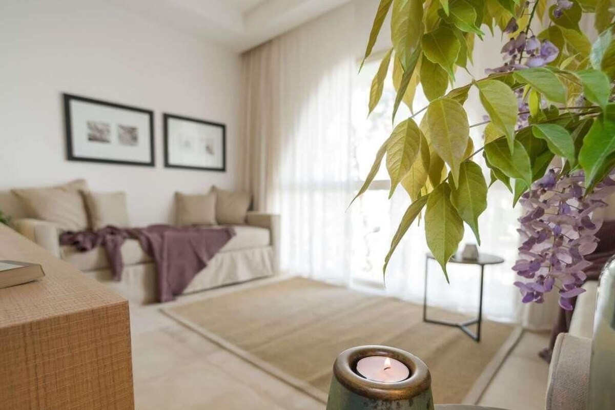 Een woonkamer met een bank, een klein tafeltje met een kaars en een plant met paarse bloemen.