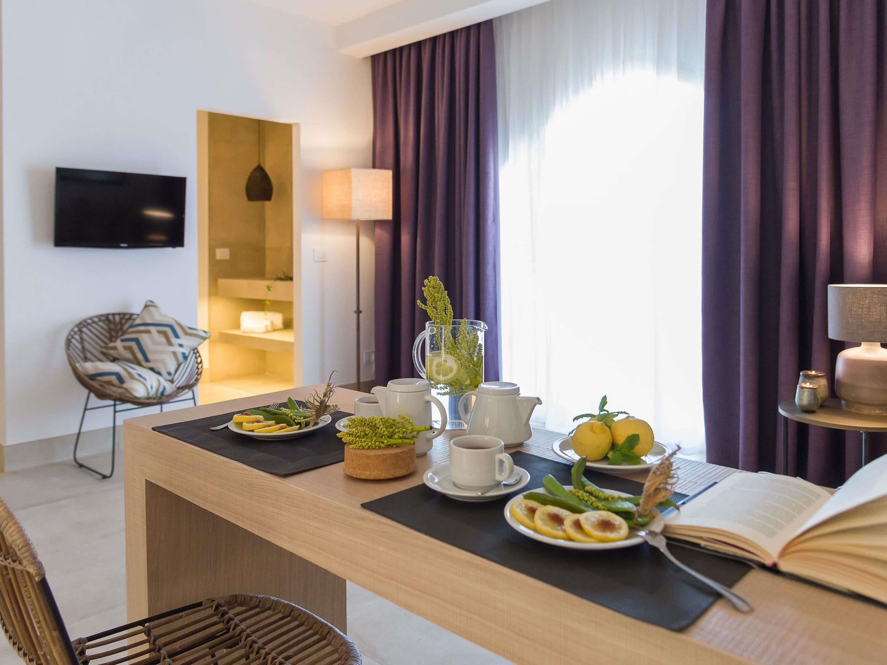 A modern hotel room with a dining area featuring a table set with breakfast, a TV, a lamp, and a chair.