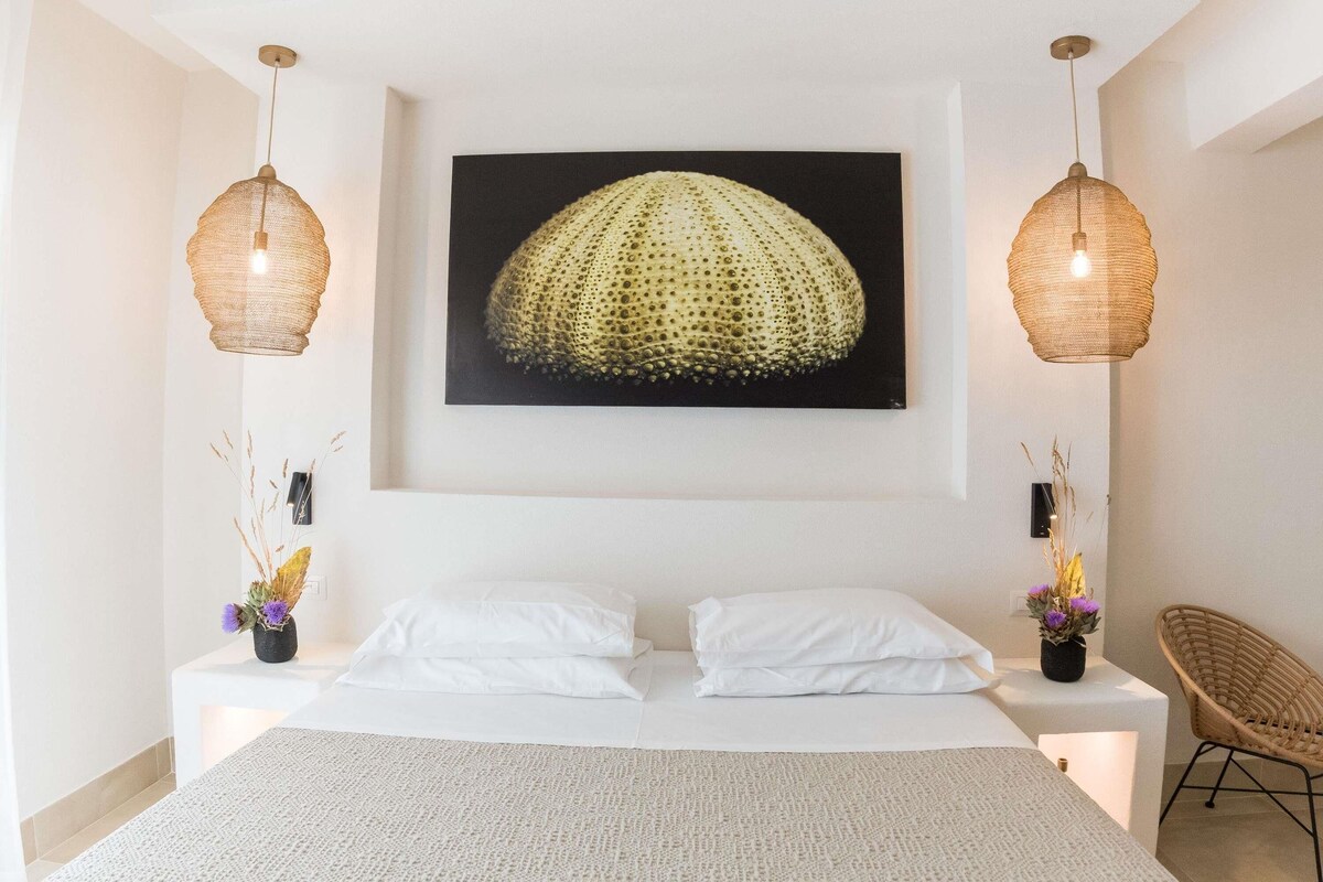 Un dormitorio con una cama, dos mesitas de noche, una silla, una obra de arte en la pared y dos lámparas colgantes.