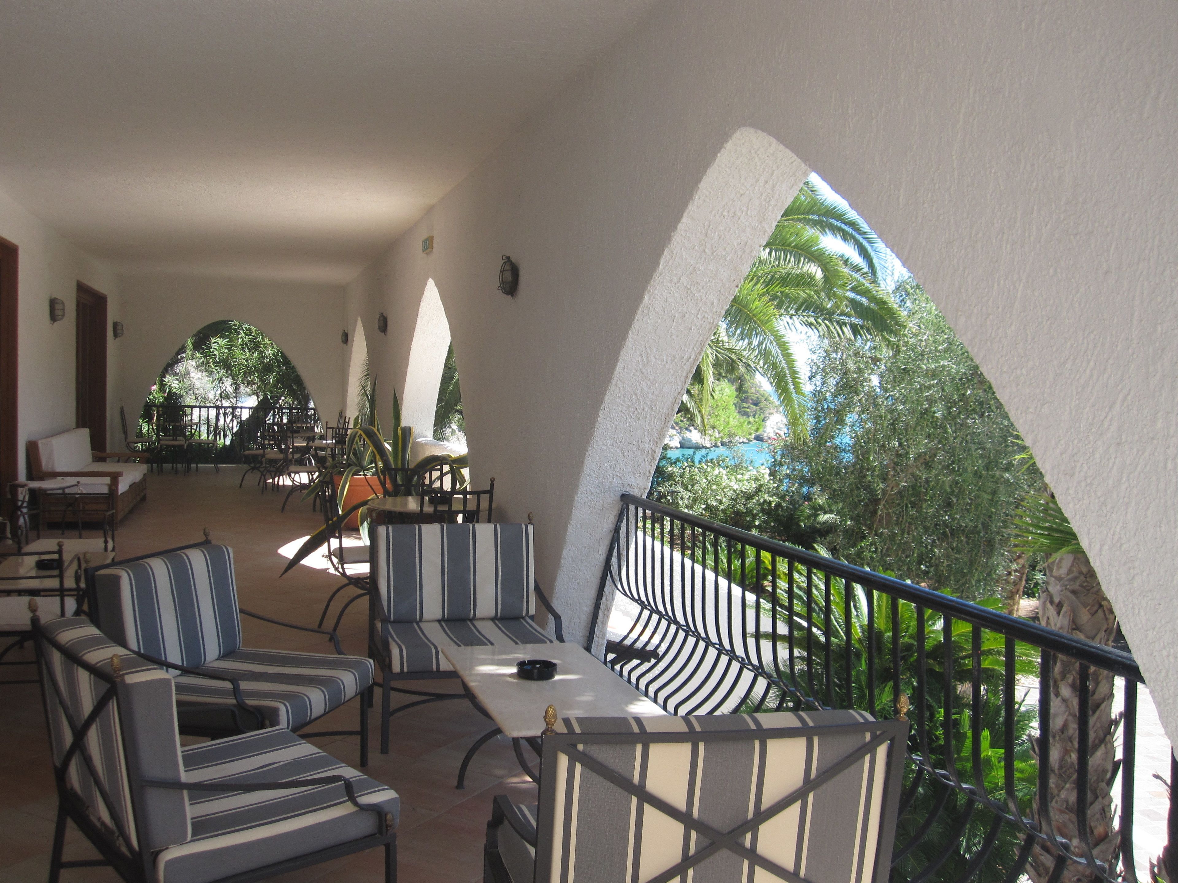 A spacious outdoor seating area with striped chairs and tables, overlooking a lush tropical garden and a clear blue pool.