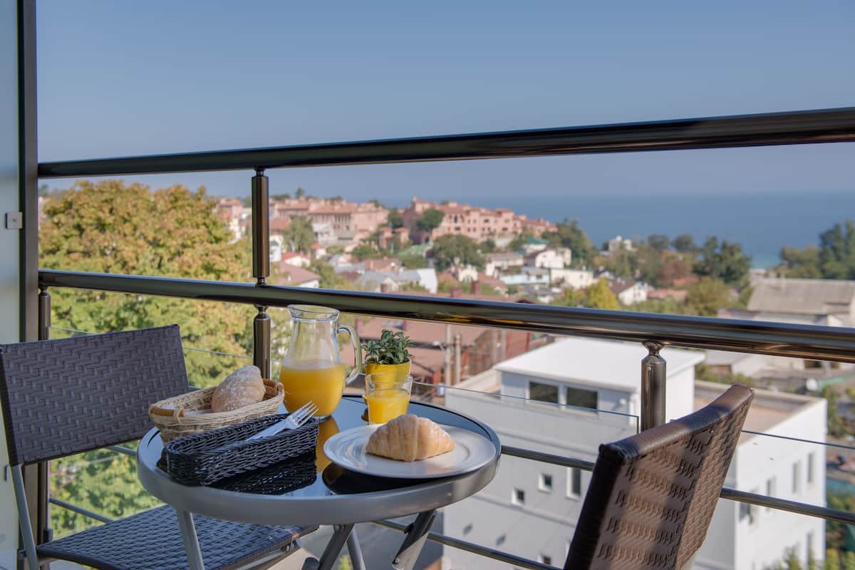 A balcony table with a croissant, juice, and bread basket, overlooking a coastal town.