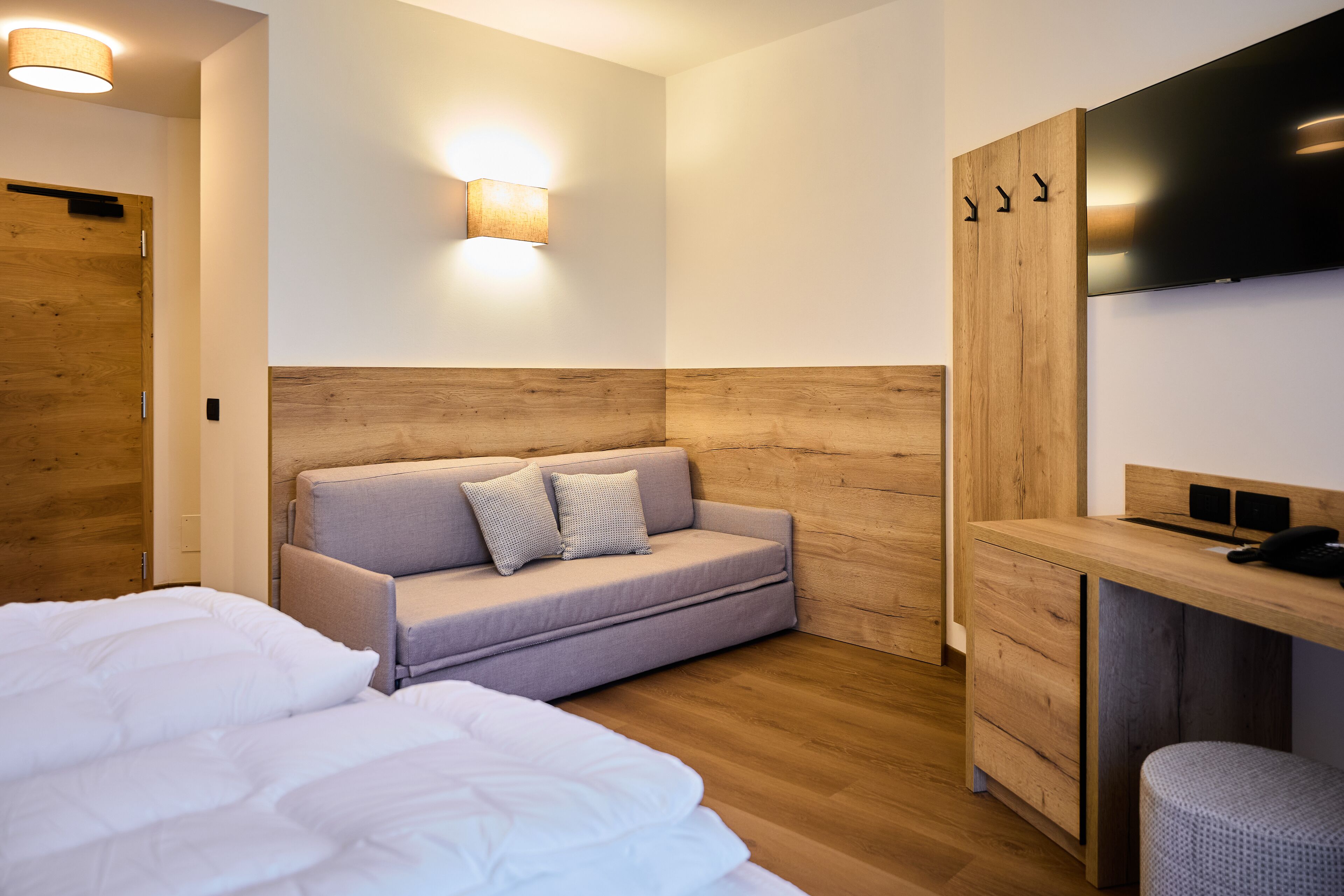 A modern hotel room with a wooden headboard, a sofa with pillows, a bed with white bedding, a desk with a chair, and a flat-screen TV mounted on the wall.