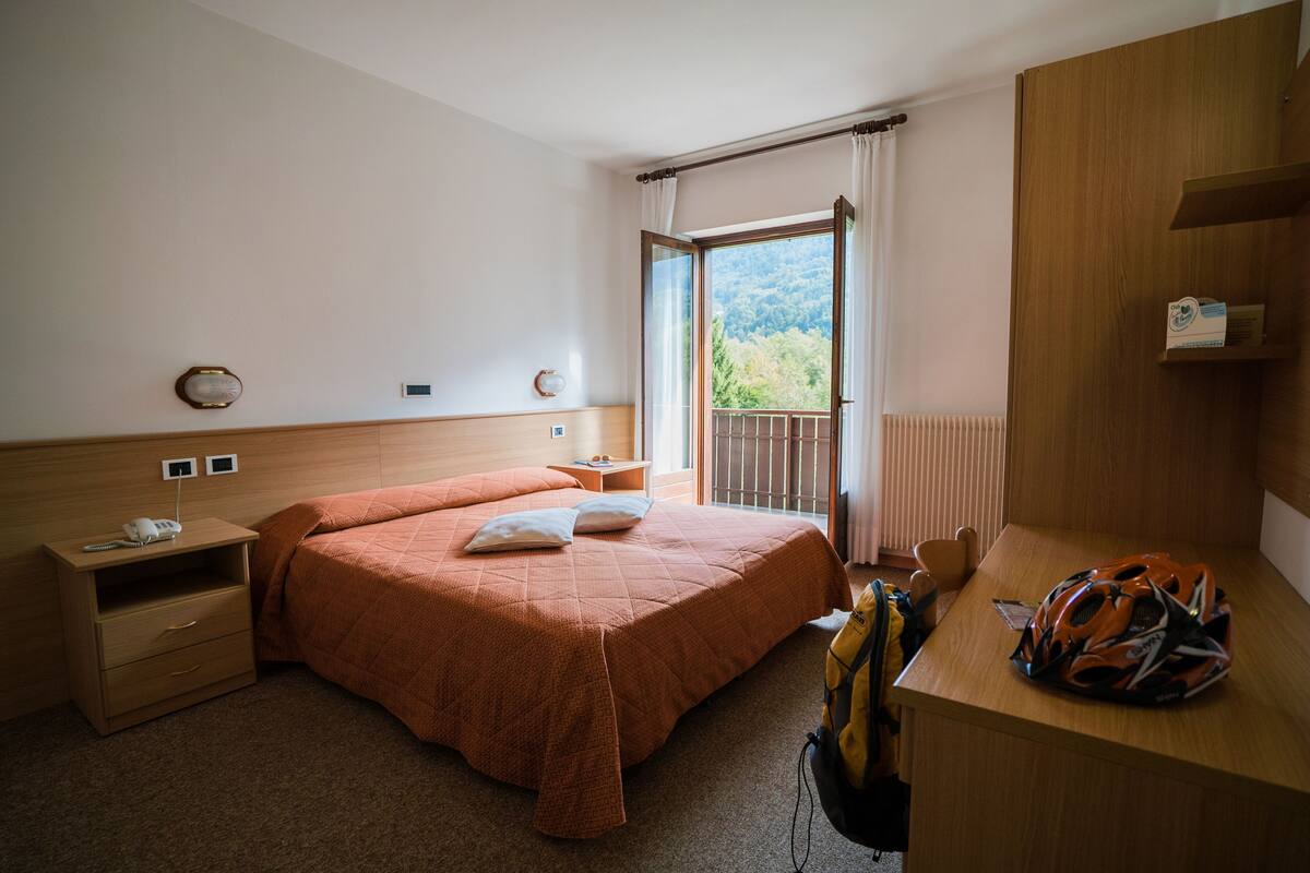 A hotel room with a bed, bedside table, and a desk with a backpack.