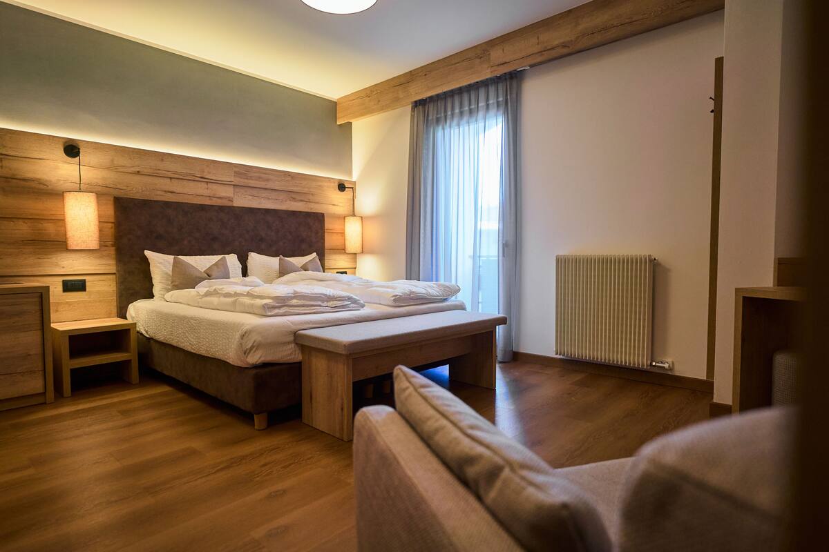 A modern hotel room with a wooden headboard, a large bed with pillows, a bedside table, a bench, and a window with curtains.