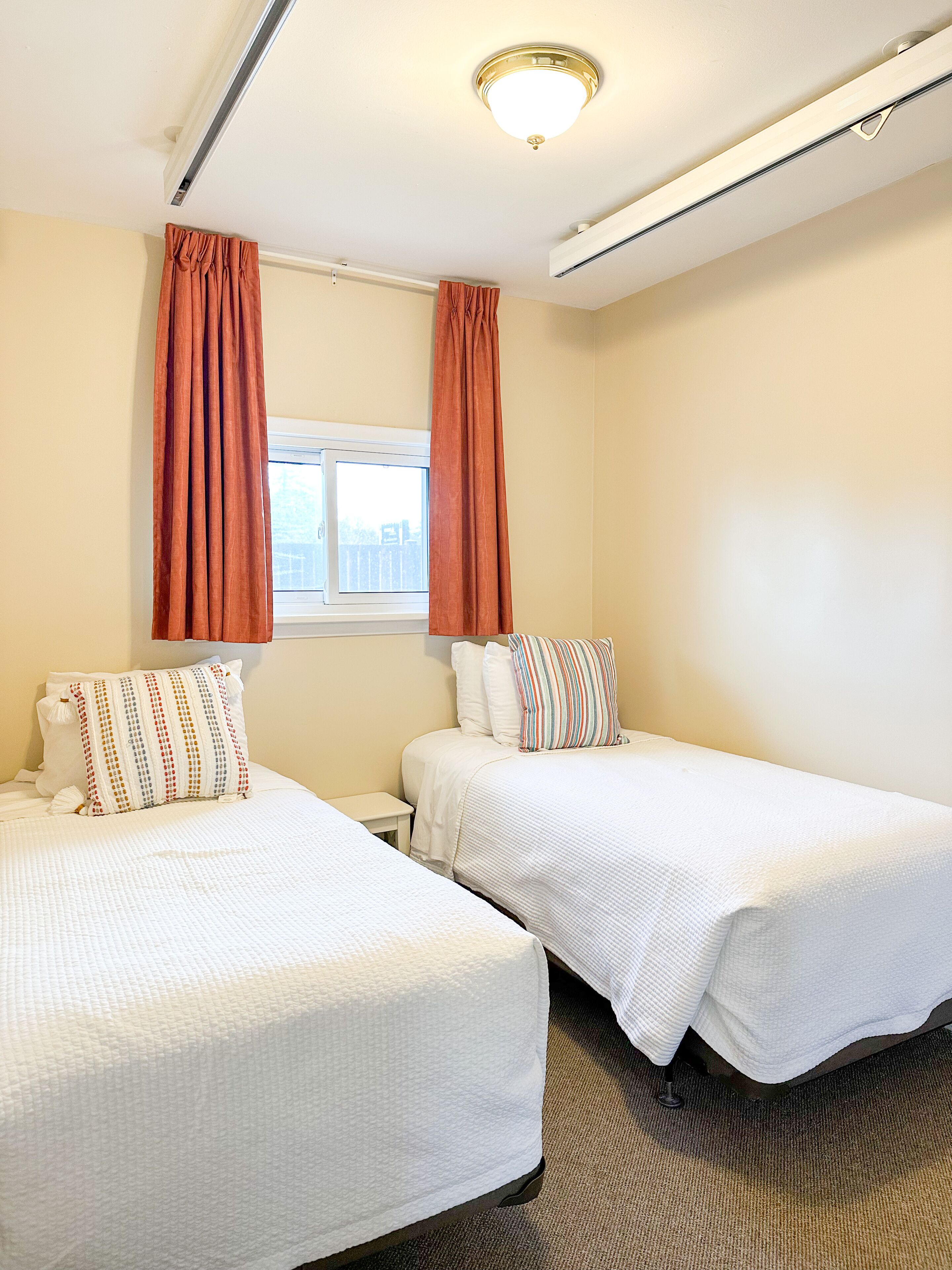 Une chambre d’hôtel avec deux lits simples, une fenêtre avec des rideaux, un luminaire au plafond et un climatiseur fixé au mur.