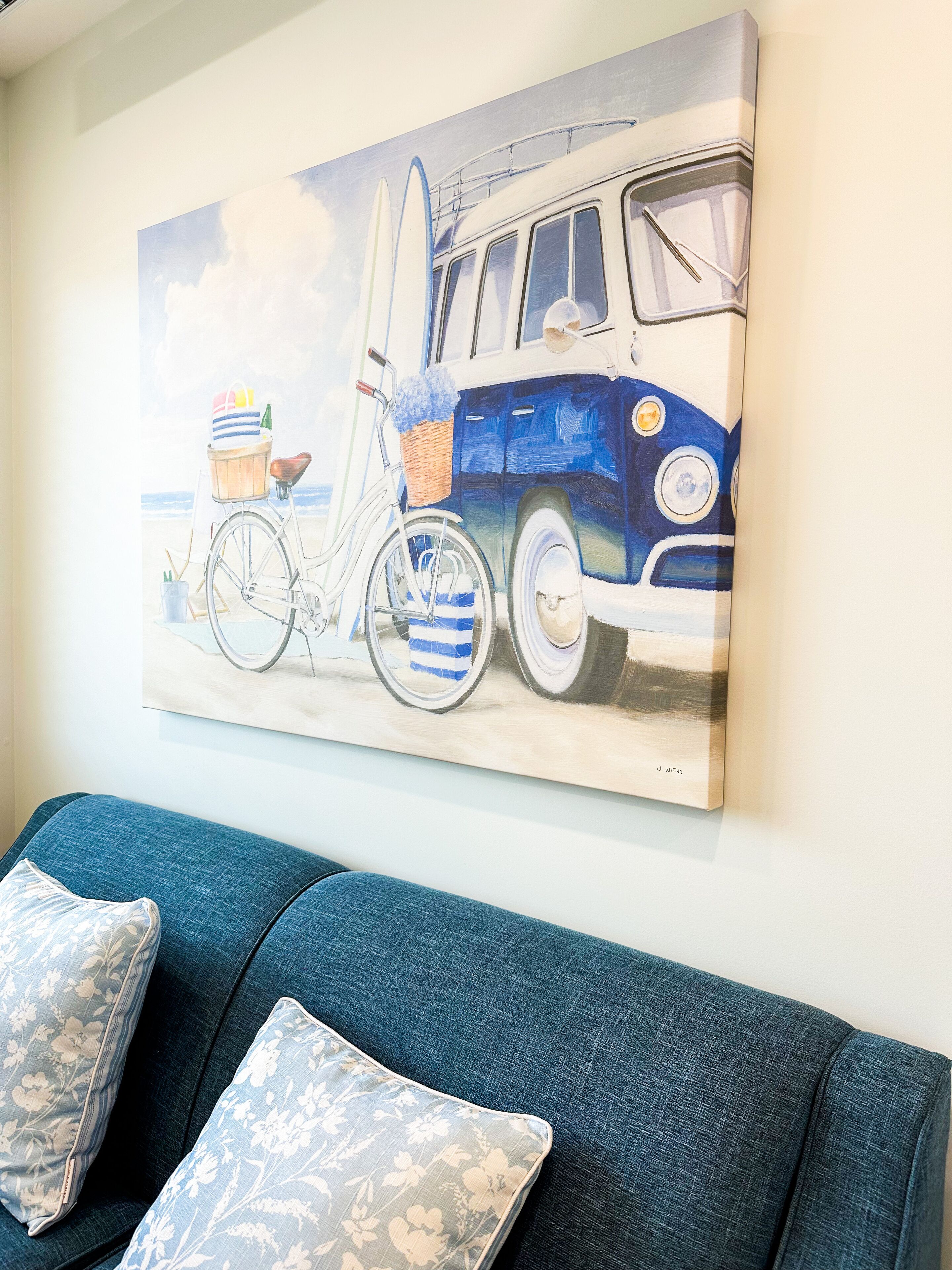 Une peinture représentant un vieux camion et un vélo sur un mur, avec un canapé bleu et des coussins à motifs floraux en dessous.