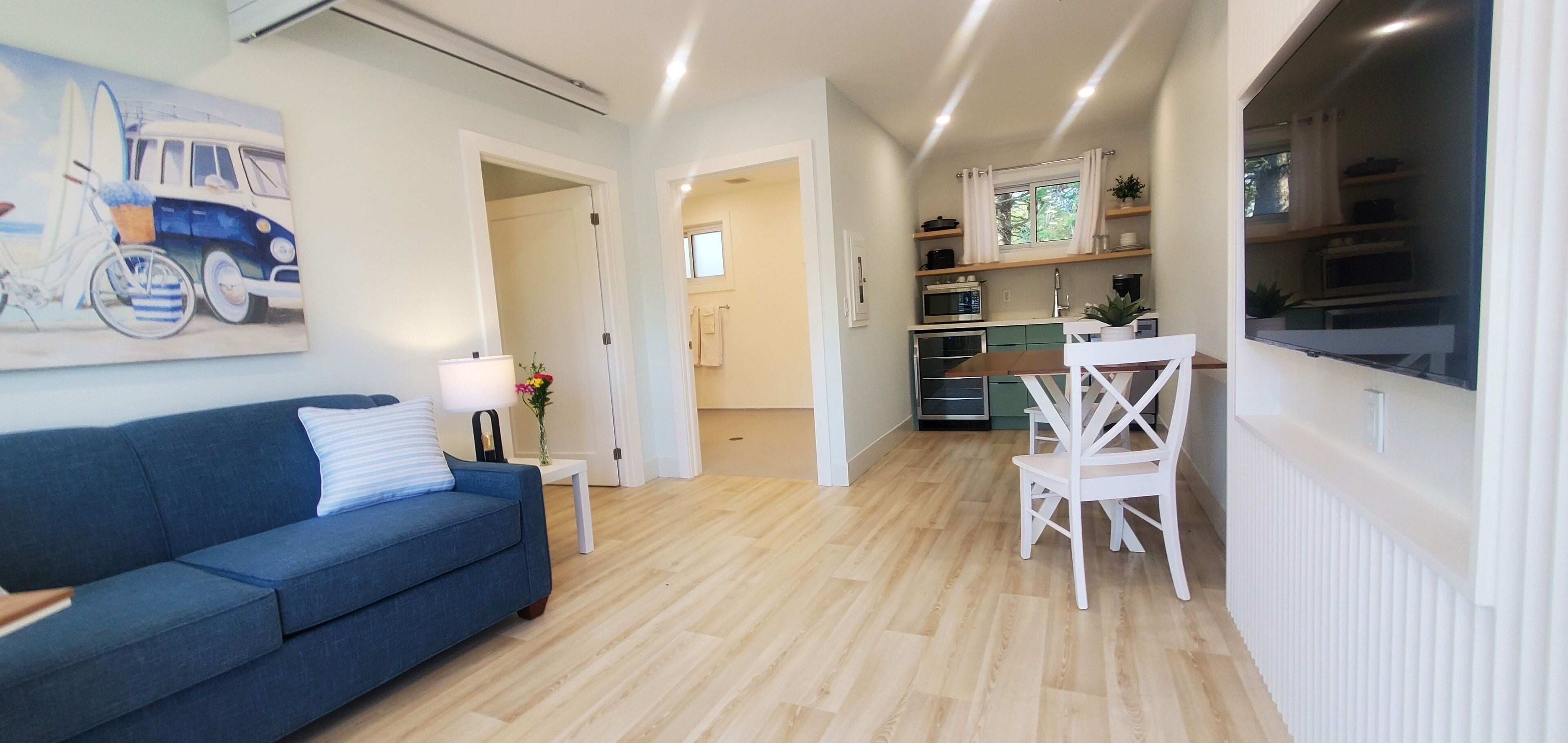 A modern living room with a blue sofa, a white dining table with chairs, a kitchen area with a microwave and cabinets, and a large window with curtains.
