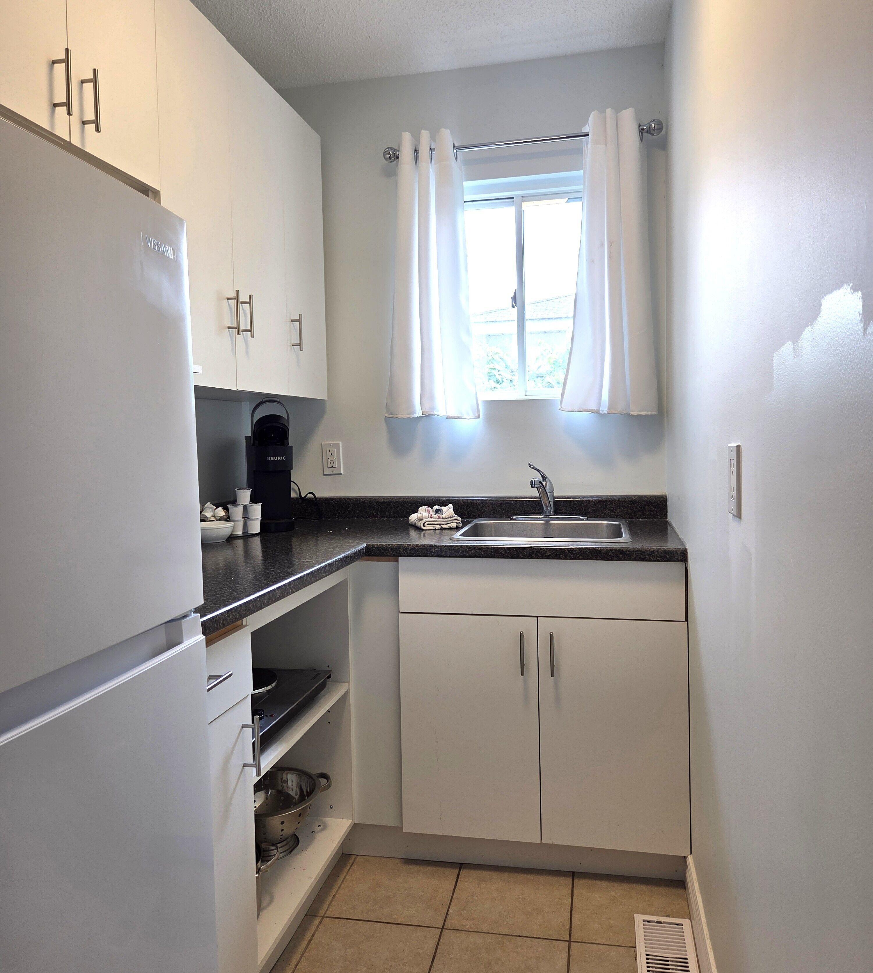 A compact kitchen with white cabinets, a built-in coffee maker, a sink, and a window with curtains.