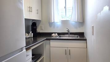 A compact kitchen with white cabinets, a built-in coffee maker, a sink, and a window with curtains.
