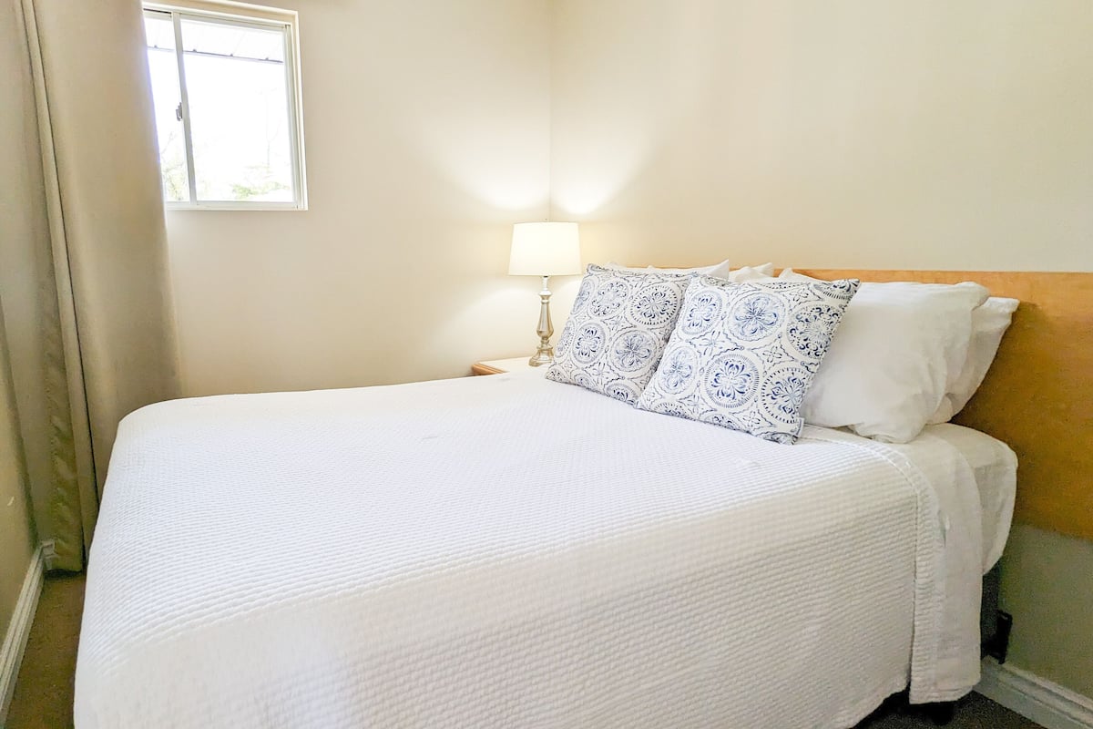 A neatly made bed with a white bedspread and two patterned pillows, a bedside lamp, and a window with curtains.